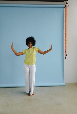 a smiling black woman stands in front of a blue photographer’s backdrop with her arms wide