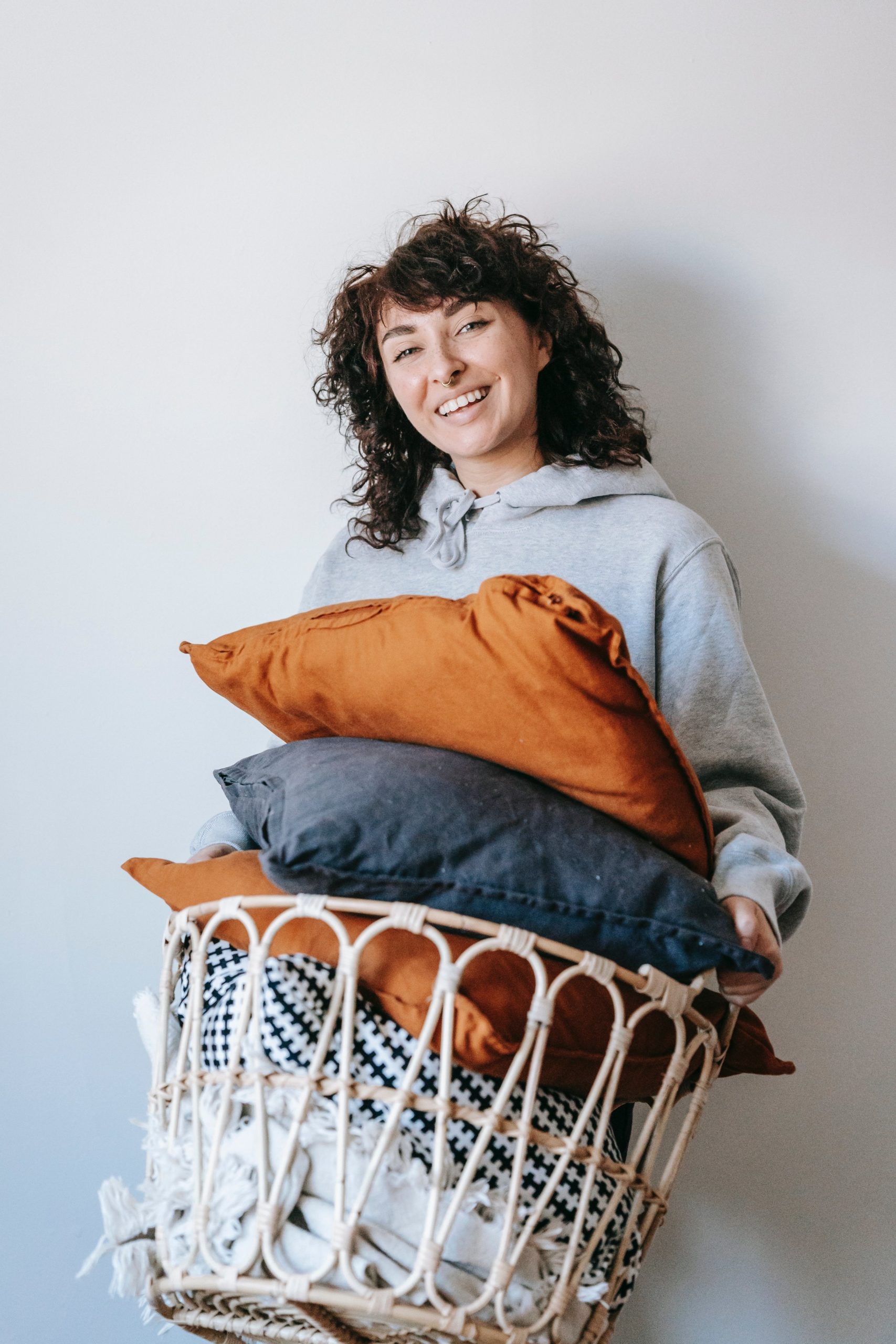 a woman in a sweatshirt carries a basket filled with linens and pillows