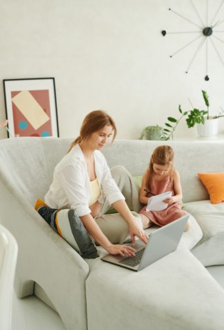 woman sitting on sofa using a computer while a young child sits next to her drawing on paper