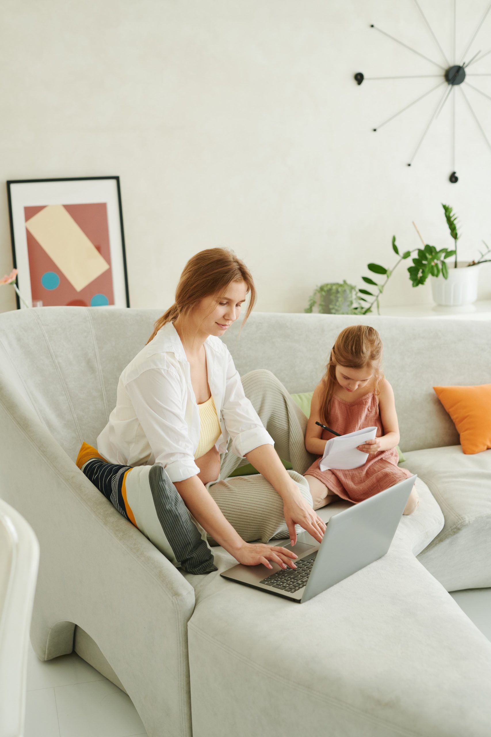 woman sitting on sofa using a computer while a young child sits next to her drawing on paper