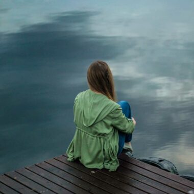 Woman sitting on a dock facing away from the camera looking out over a lake.