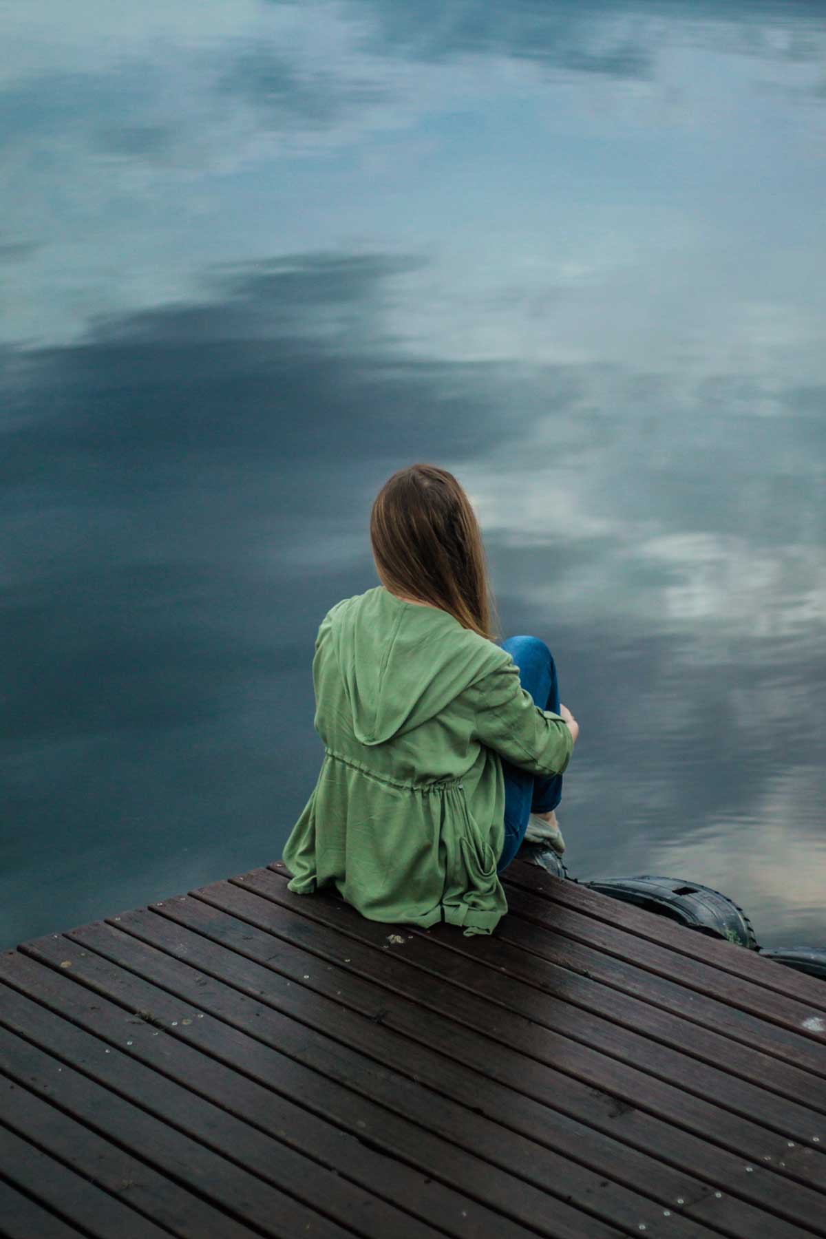 Woman sitting on a dock facing away from the camera looking out over a lake.
